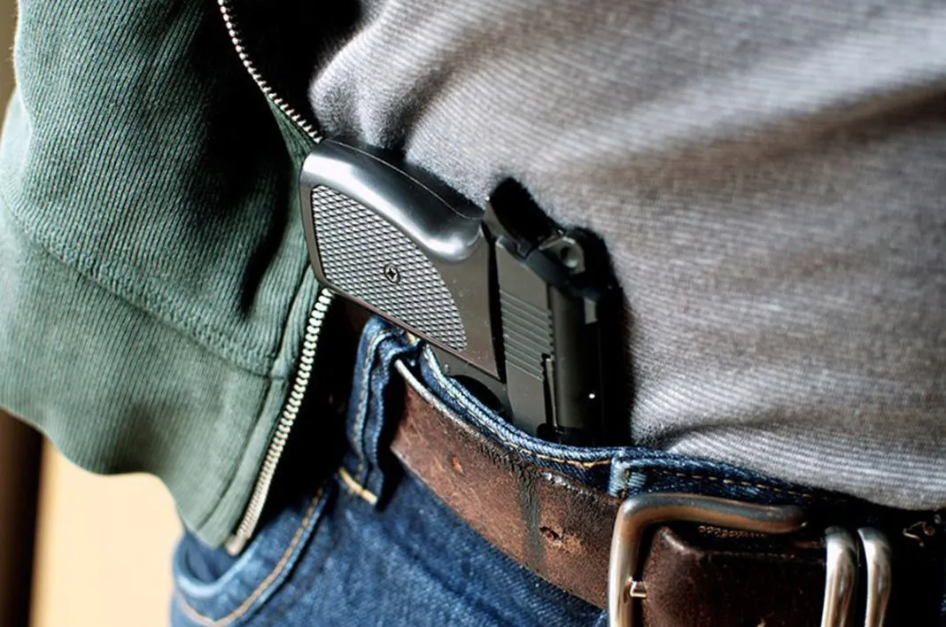 Uma pessoa, com camisa cinza, calça jeans e cinto marrom carrega uma arma preta na cintura