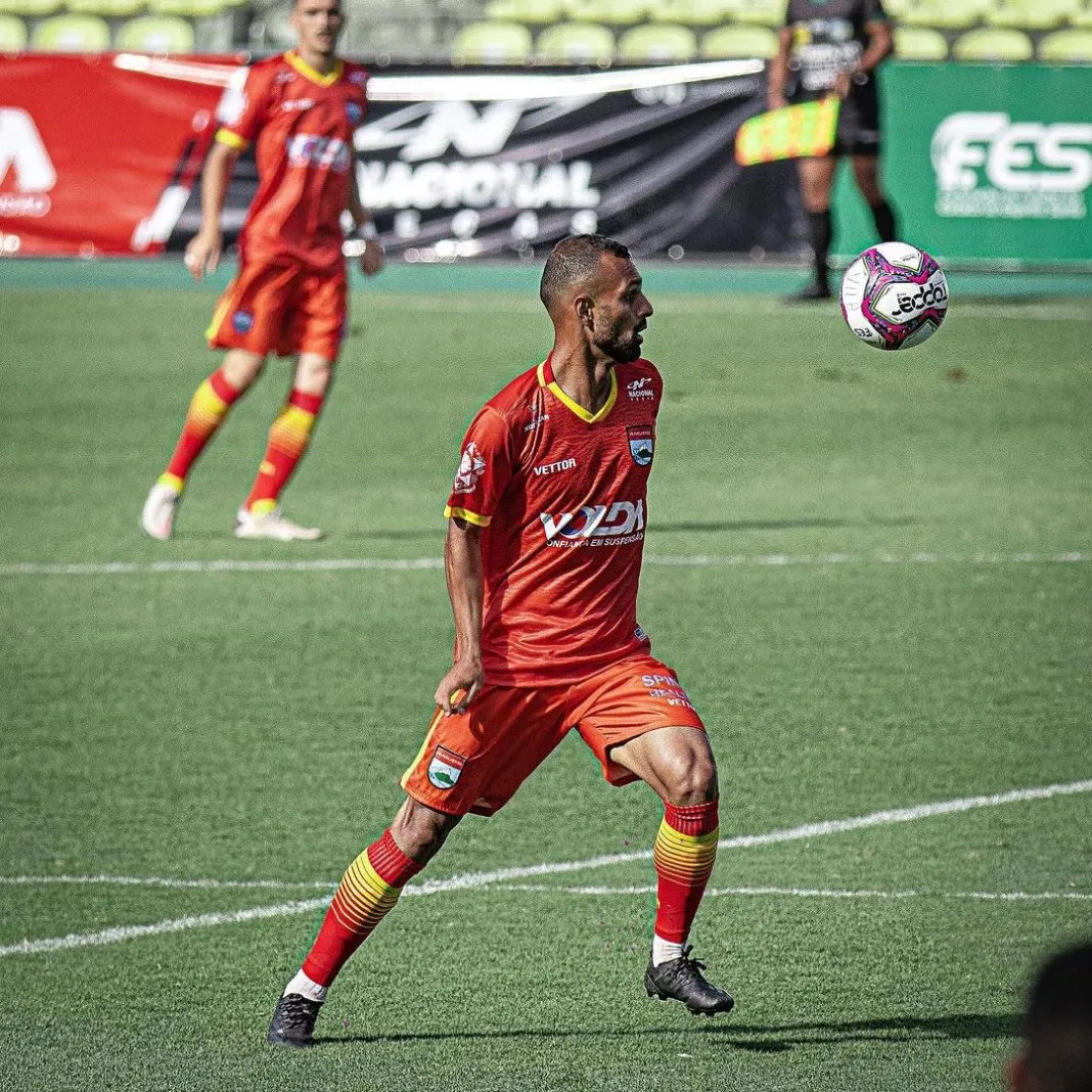 Um homem com uniforme (camisa e calção) laranja corre em um campo, no gramado verde. Ele joga bola.