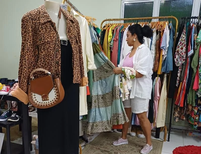 Em primeiro plano, um manequim vestido com roupas de um dos brechós do Projeto Circulando; ao fundo, interessada avalia vestido pendurado em arara de roupas