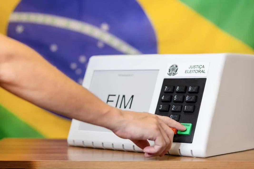Urna eletrônica com a bandeira do Brasil ao fundo.